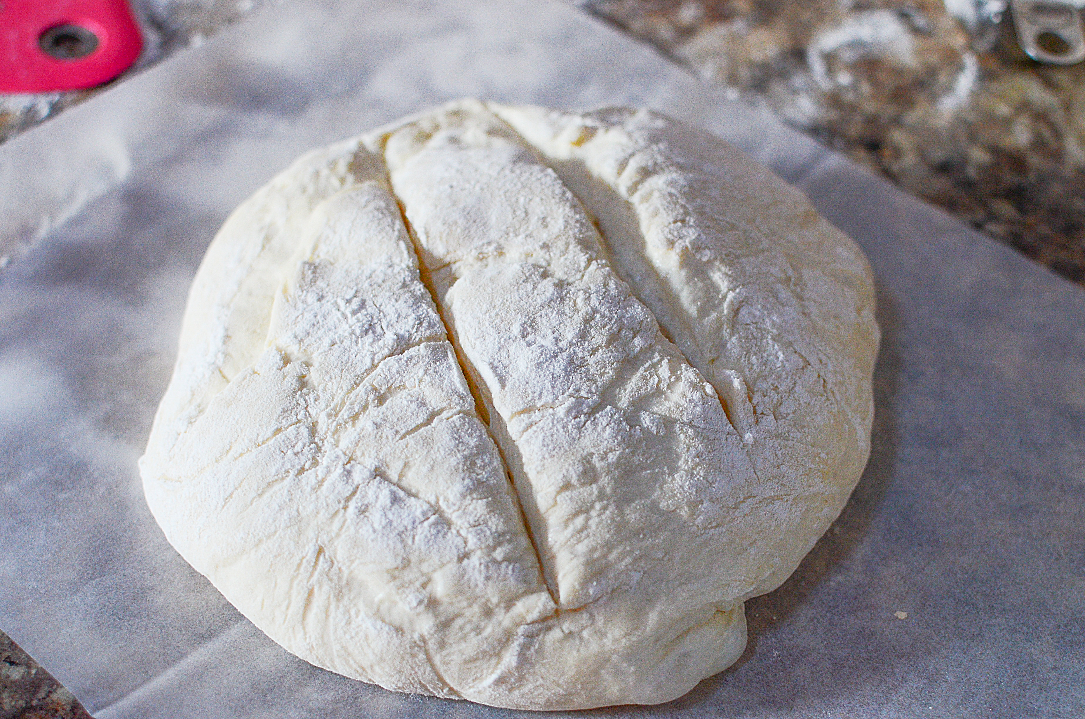 A round ball of bread dough sitting on parchment paper with two diagnal cuts in the top