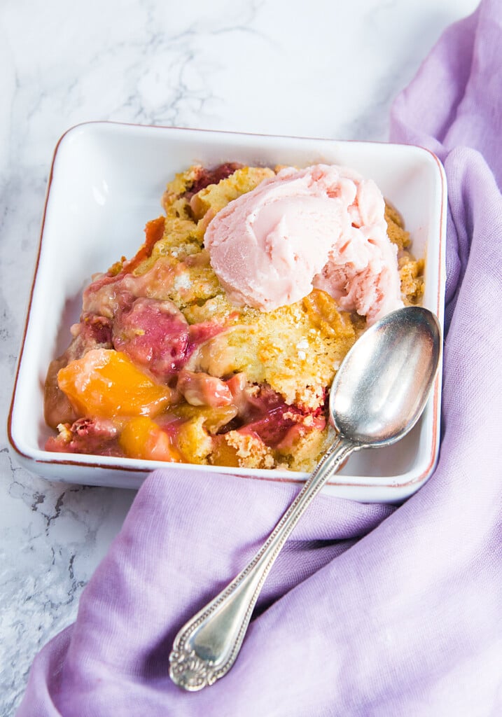 Fruit cobbler in a square white bowl with a spoon resting on the side.