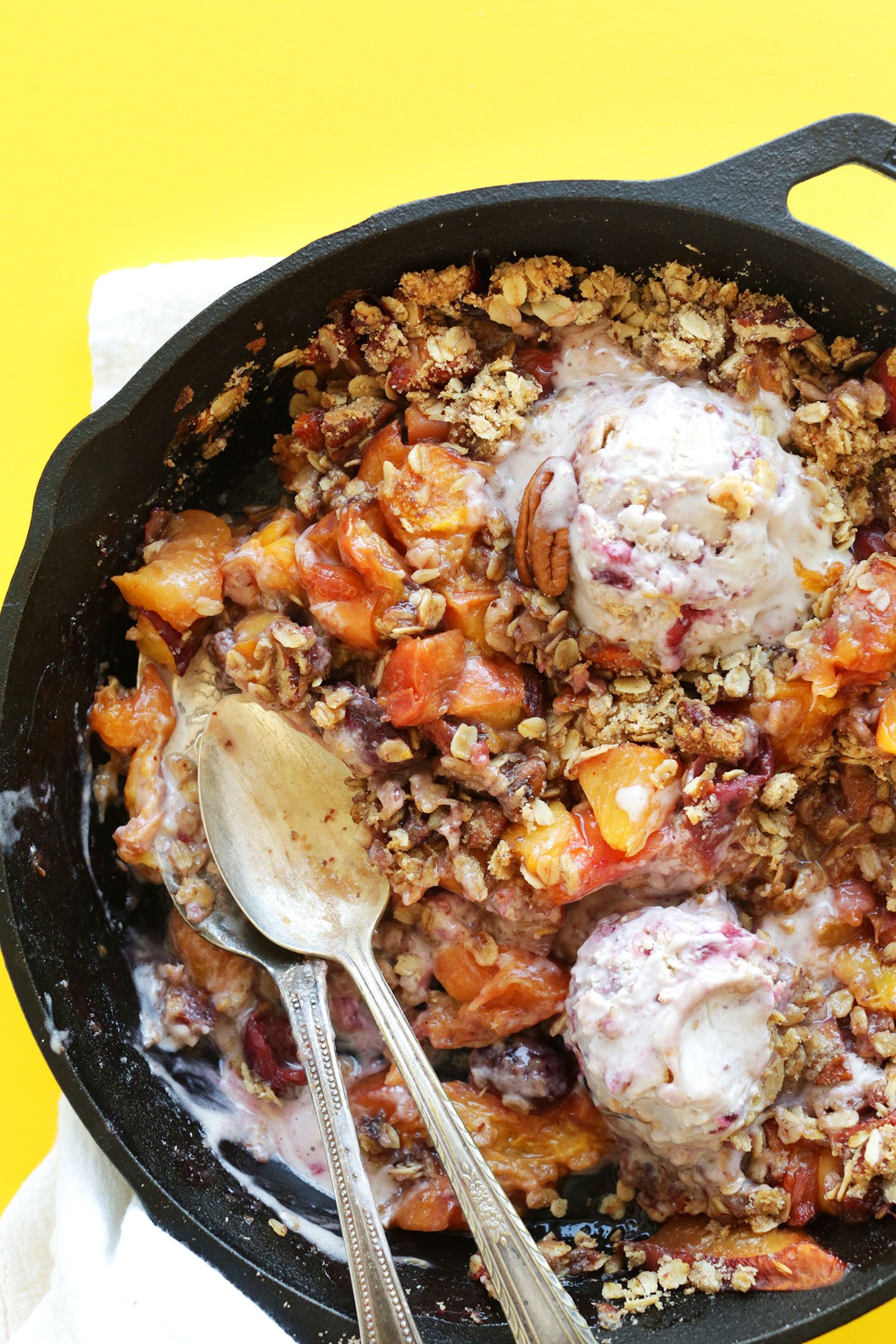 Peaches, ice cream and a crisp topping all inside a cast iron pan with two spoons. 