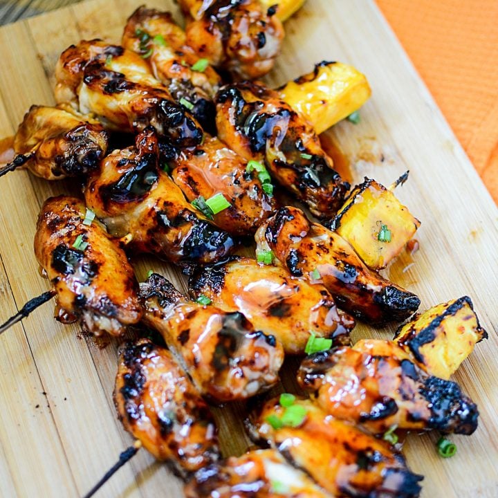 Five wings per skewer with a pineapple on the end, sit on a wooden cutting board. There are 4 skewers in the photo.