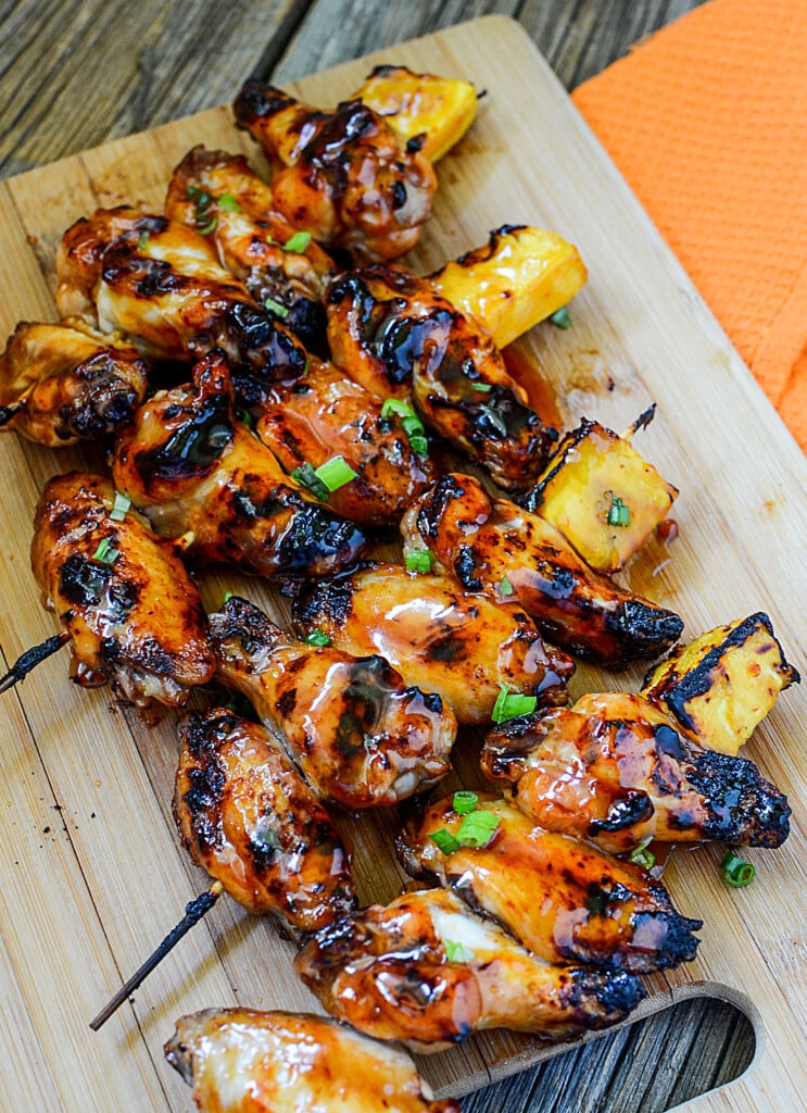 Skewers of grilled chicken wings on a cutting board with pineapple on the ends.