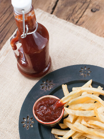 Ketchup in a bottle with fries on a plate. A small amount of ketchup is in a bowl on the plate as well.