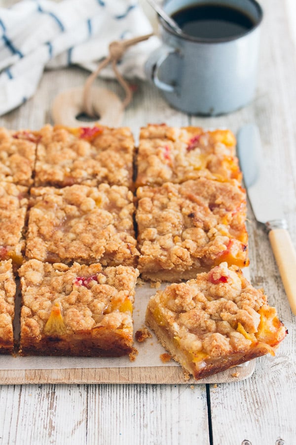 Peach crumb bars on a wooden plate with one bar sitting askew to the others. A knife sits beside them with a blue coffee up on the background.