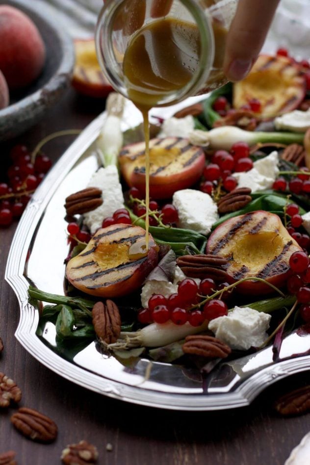 Grilled peaches on a silver plate with pecans, greens and cheese. A dressing is being poured over top the peaches.
