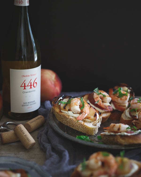 Grilled shrimp and peach crostini on a dark plate with a bottle of wine next to it.
