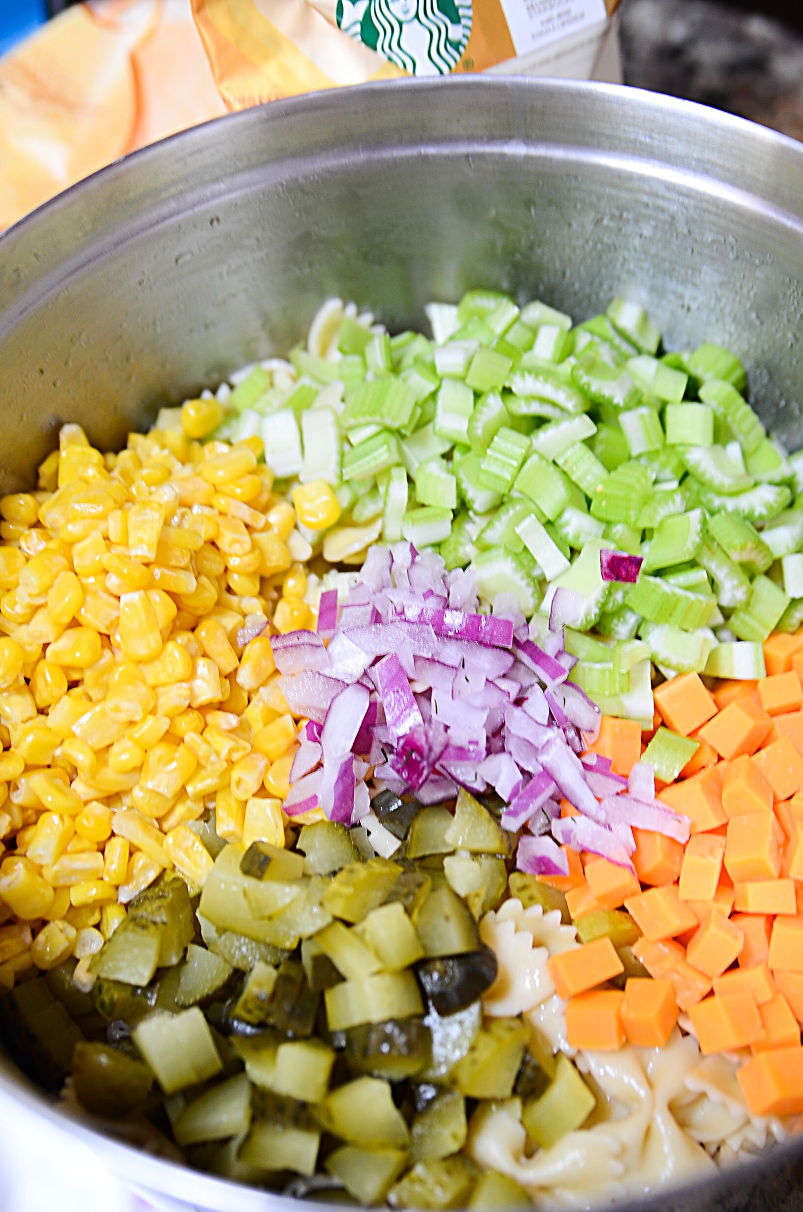 Corn, celery, cheddar cheese, pickles and red onions sit on top of cooled pasta in a silver bowl. 