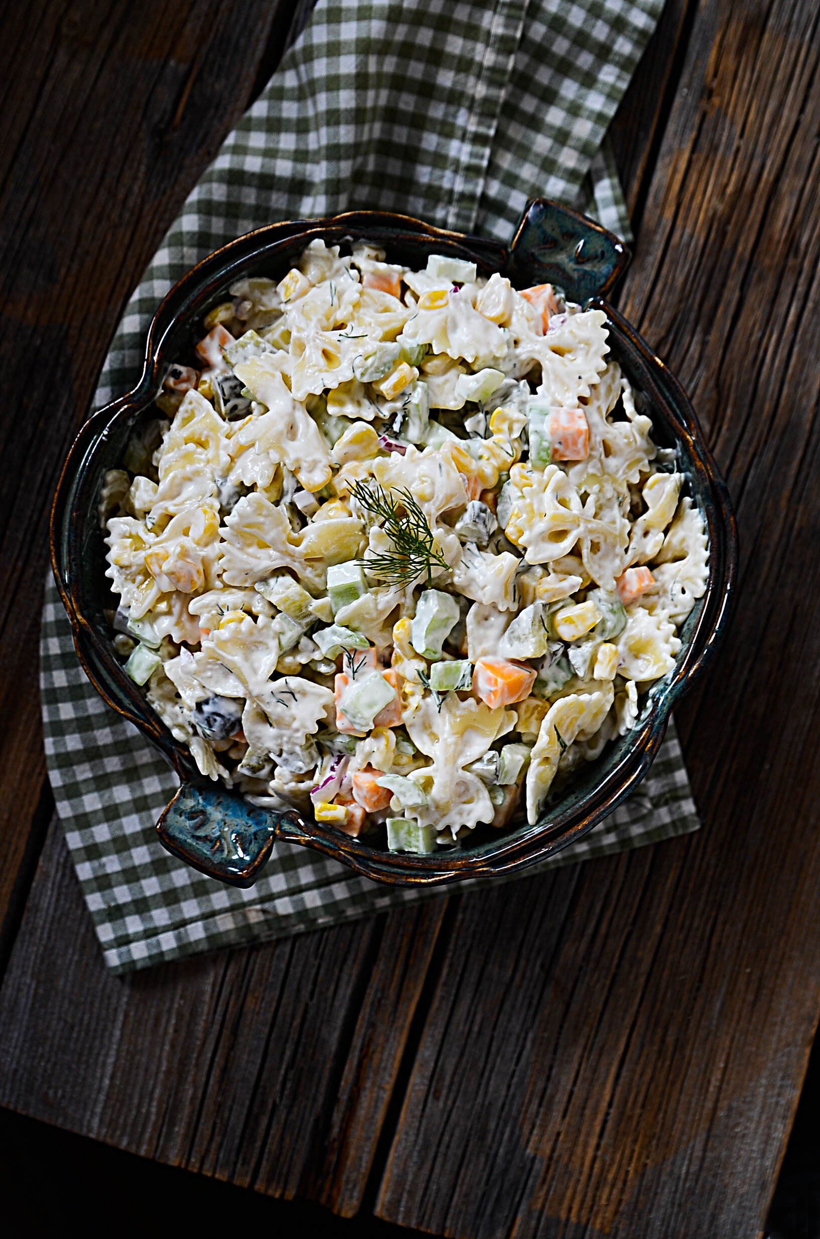 Creamy dill pickle salad with pickles in a teal pottery bowl. The bowl sits on a checkered napkin on a wood table. 