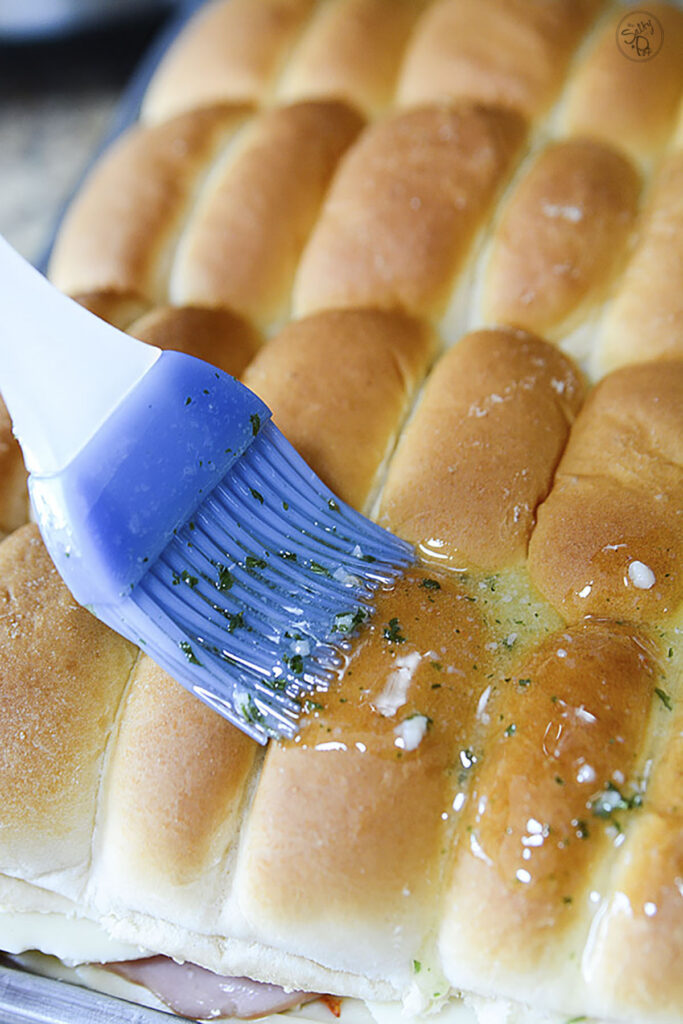 Garlic butter being spread on top of the tray buns with a blue silicone brush.