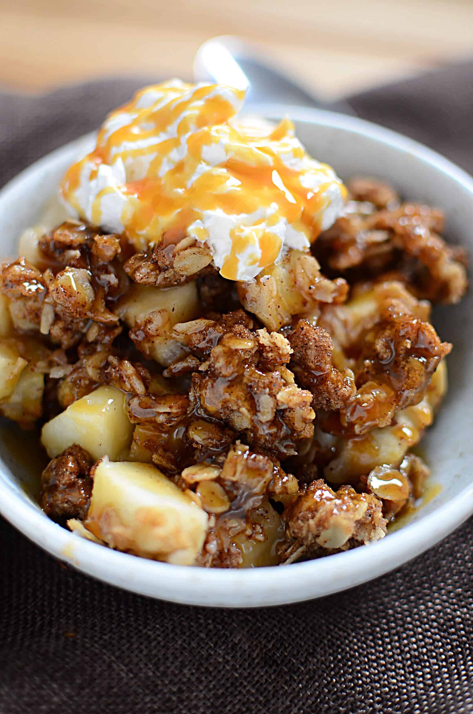 Spiced apple crisp with caramel drizzled on top in a white bowl with whipped topping