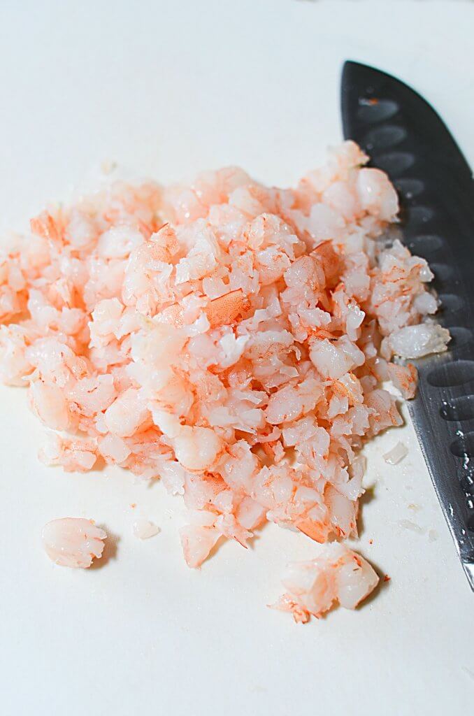 A pile of finely diced cooked shrimp sitting on a white plastic cutting board with the blade of a knife sitting next to it.