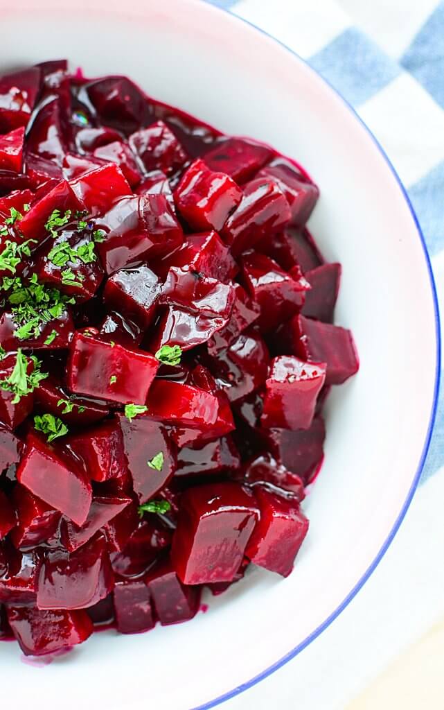 This half photo of the ruby jeweled beets are cut into bite size pieces in a sauce. The contents are sitting in a blue rimmed enamel bowl. 