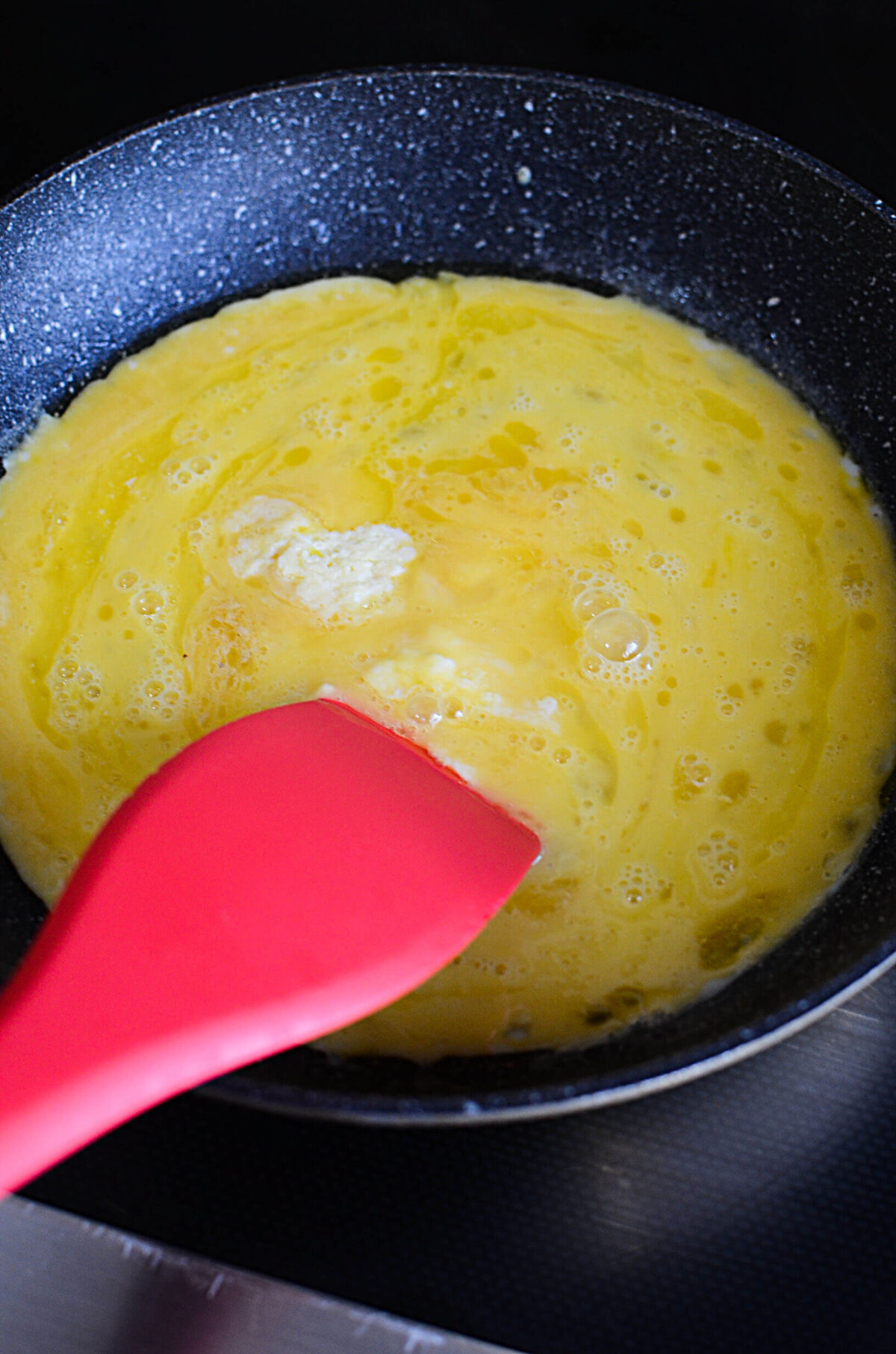 A red spatula stirs cooking eggs in a frying pan.