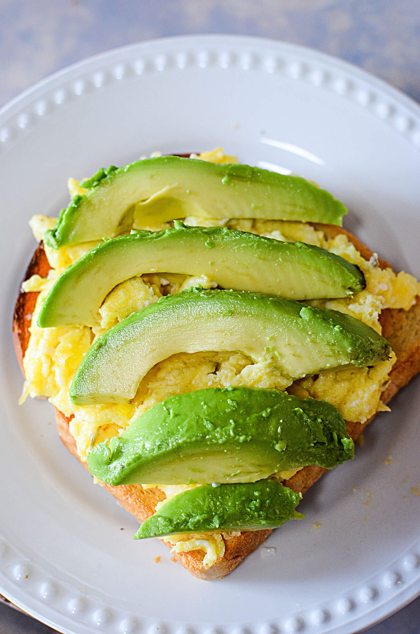 Avocado that's been sliced is sitting on top of scrambled eggs and toast on a white plate.