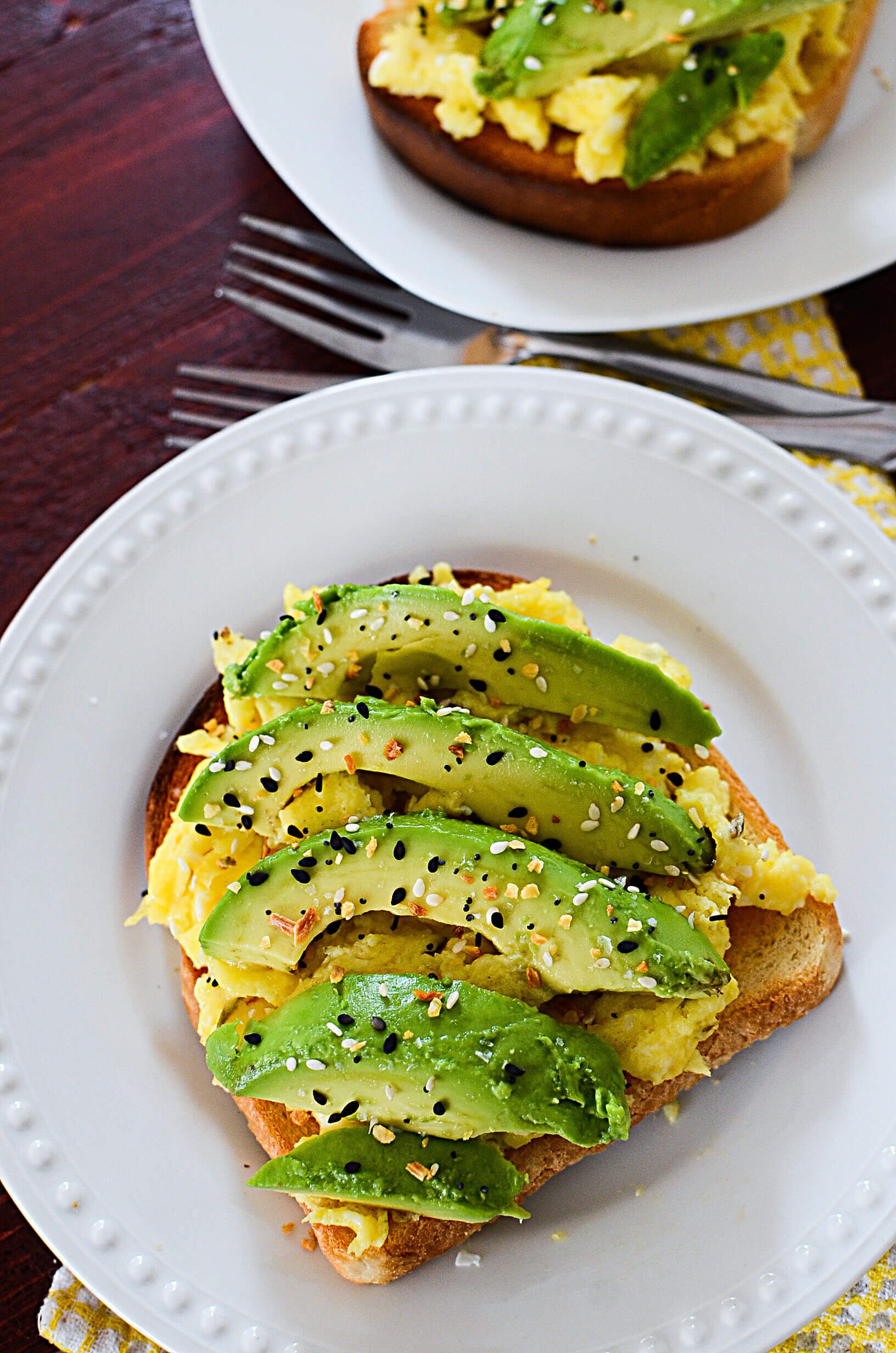 The avocado toast sitting on a white plate with two forks above it. There is another serving of toast above the forks.