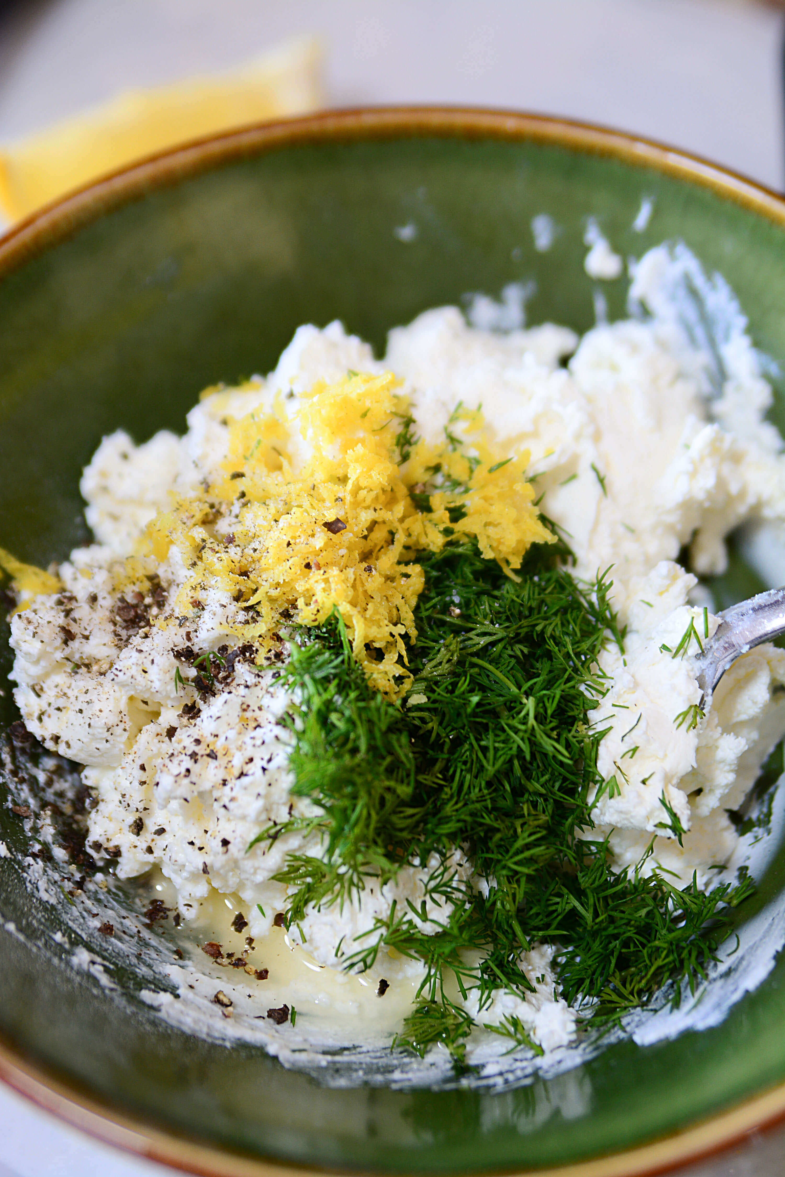 Lemon, dill, salt and pepper being added to the cheese in a green bowl.