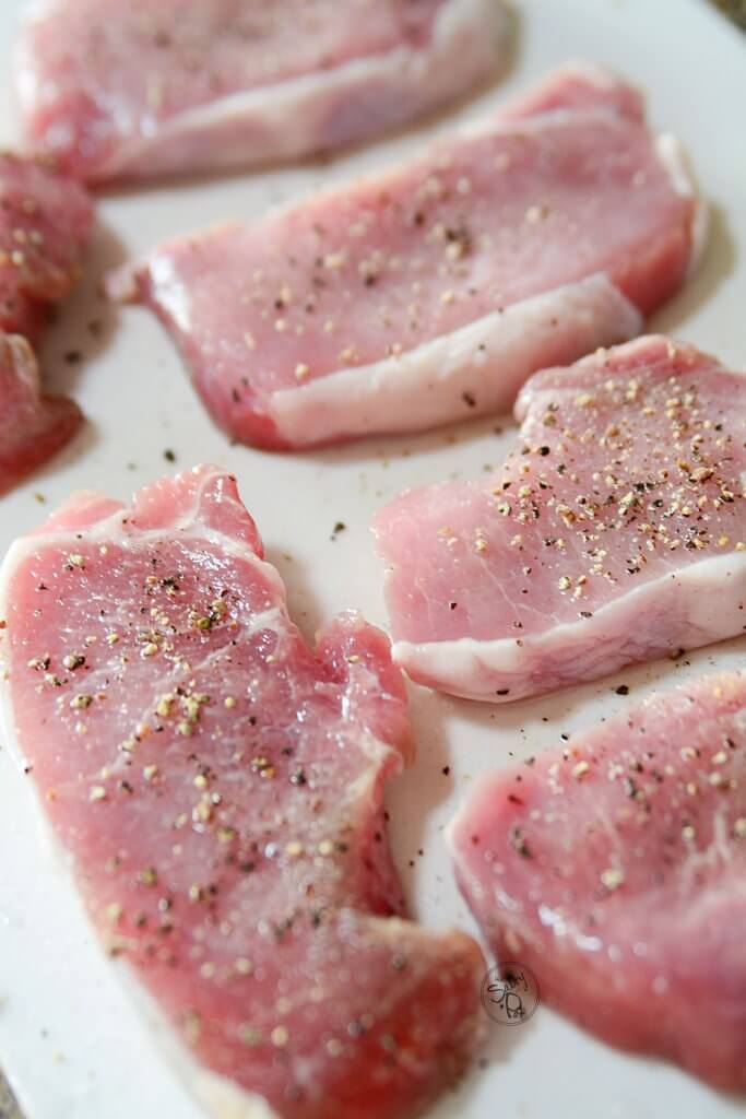 Raw pork chops on a plastic white cutting board, sprinkled with salt and pepper.