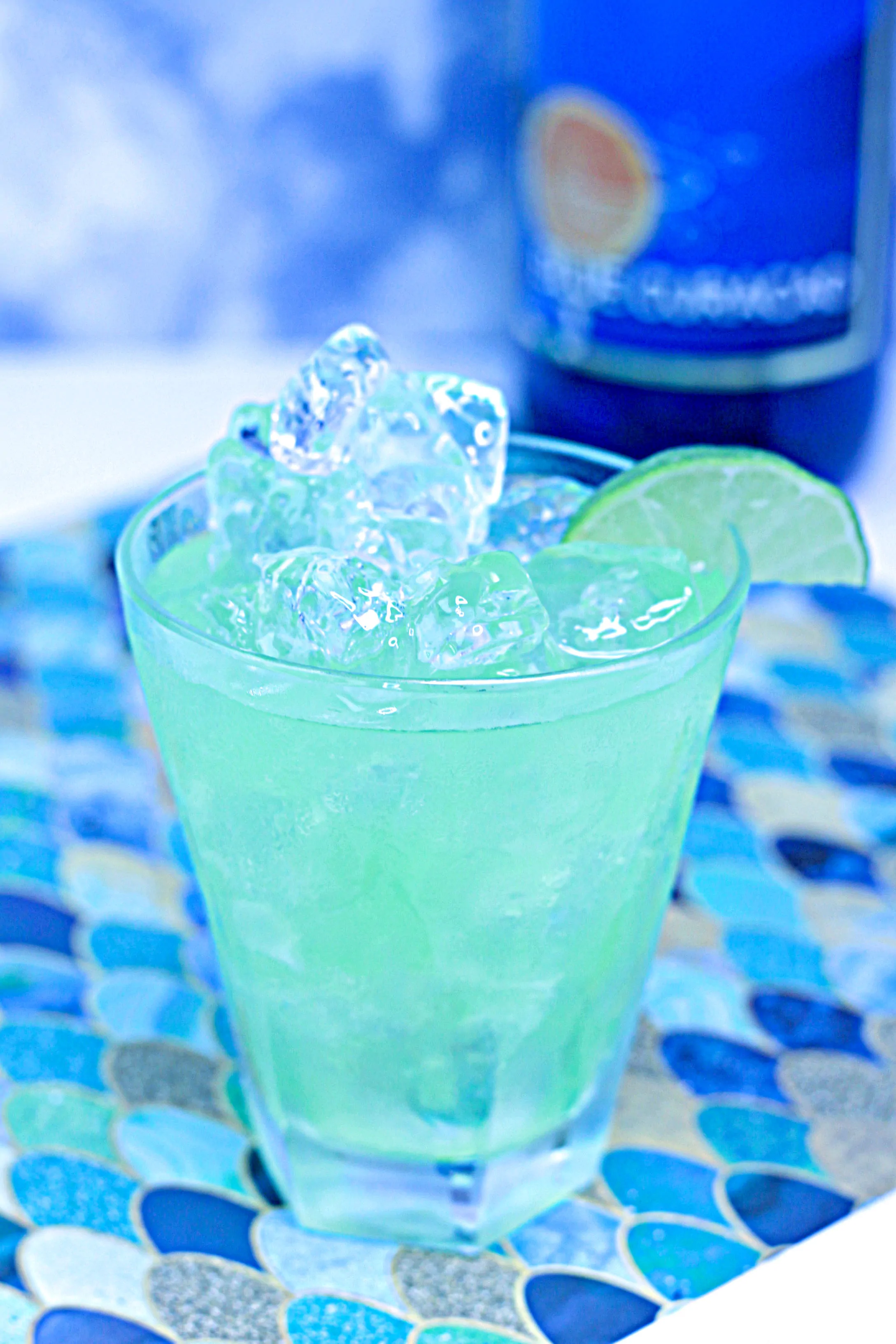 A green liquid beverage in a clear glass. A blue bottle is in the background.