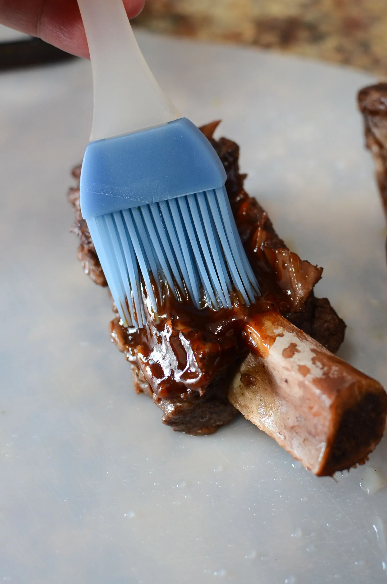 A blue silicone bbq brush, brushing bbq sauce over a beef short rib.