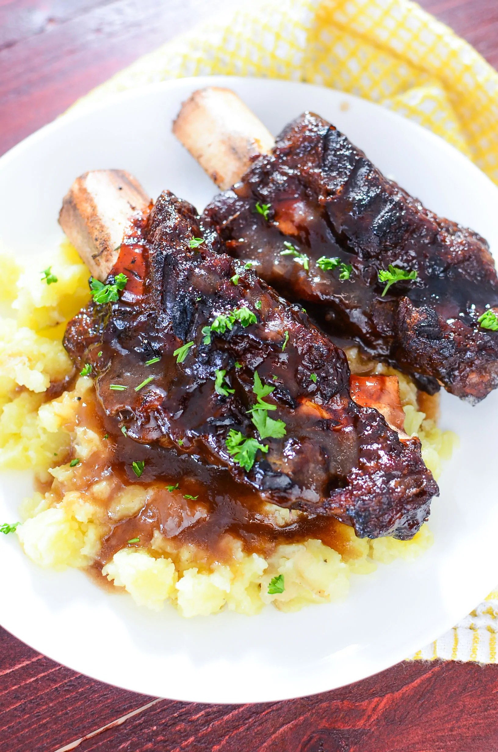 A delicious looking photo of two bbq beef short ribs resting on a bed of mashed potatoes with gravy, on a white plate.