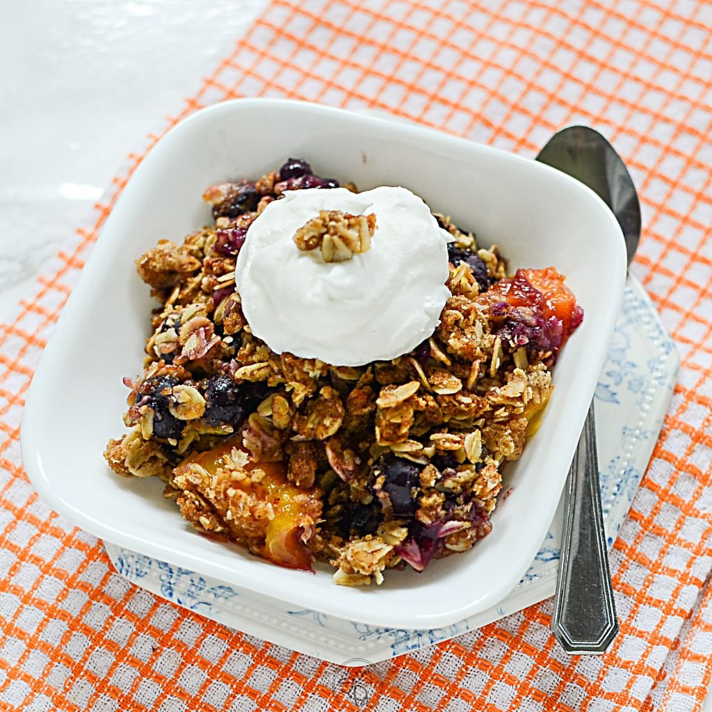 The Peach crisp is in the bowl with whipped cream on top. The handy spoon is beside the bowl that's sitting on a pretty blue plate all resting on an orange napkin.