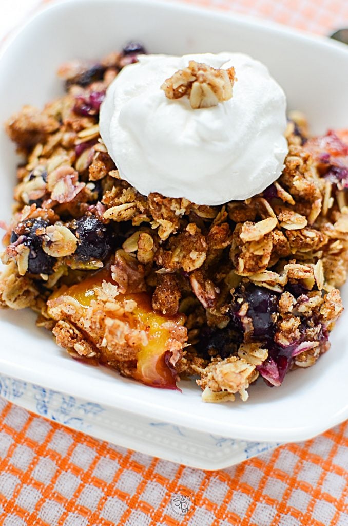 Peach and blueberry crisp in a white bowl with whipped cream on top.