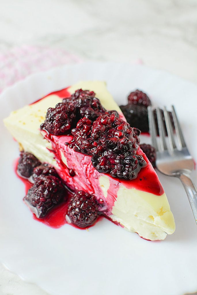 A slice of cheesecake on a white plate with some berry sauce on top. Beside it is a spoon.
