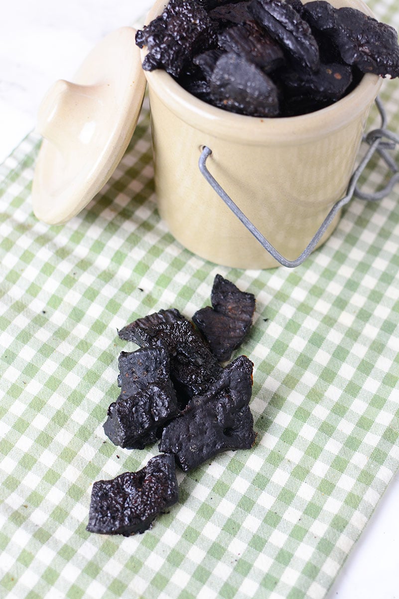 A picture of seven pieces of homemade liver dog treats sitting on a green and white checkered table cloth. Next to it is a small ceramic container full of liver treats with the lid sitting on the side of the container