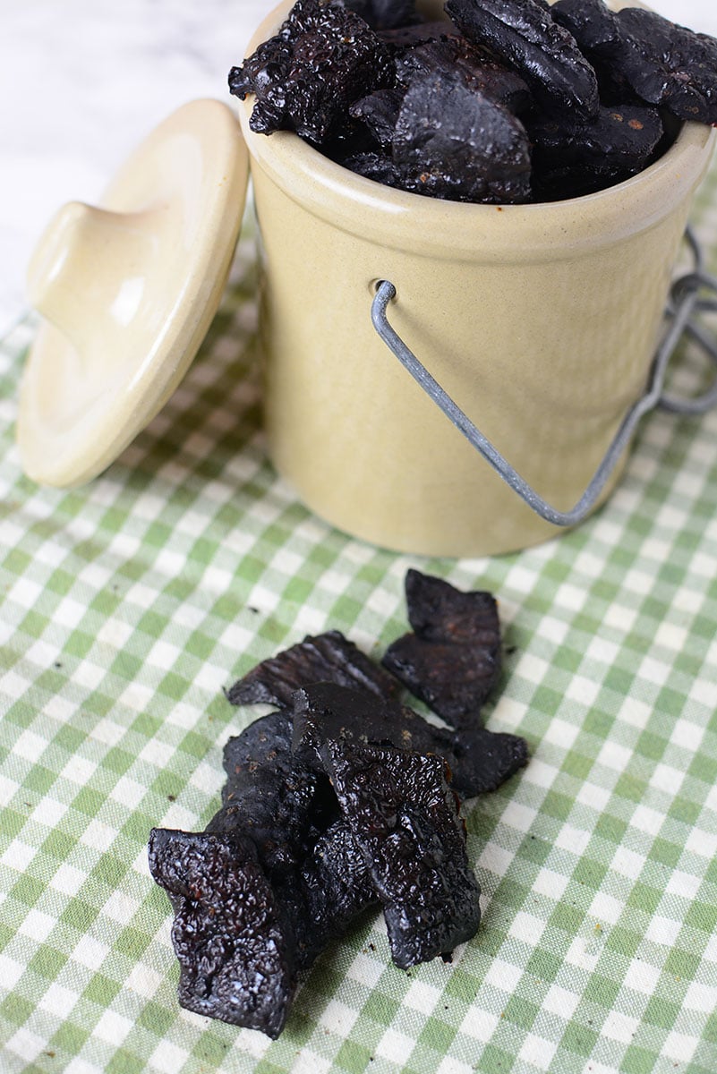 A picture of seven pieces of homemade liver dog treats sitting on a green and white checkered table cloth. Next to it is a small ceramic container full of liver treats with the lid sitting on the side of the container