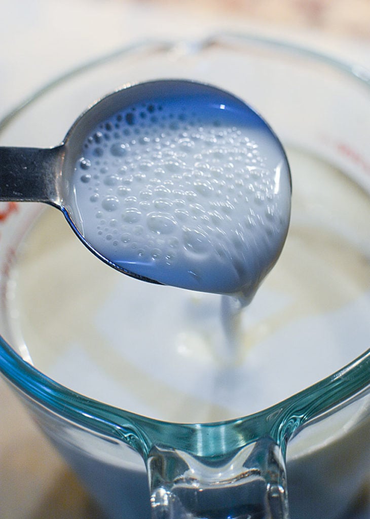 A close up image of a 1 tsp measuring spoon with some milk on it. The milk on the teaspoon is being poured into a glass measuring cup