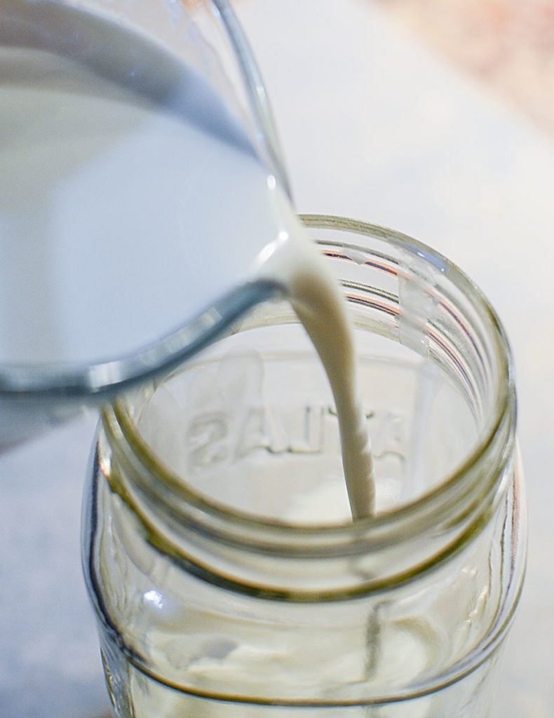 Buttermilk is being poured from a glass measuring cup into a glass jar. 