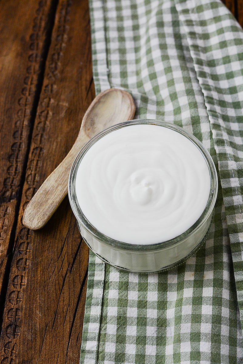 A small bowl of home made CREME FRAICHE sitting on a green and white checkered table cloth on a decorative wodden cutting board.