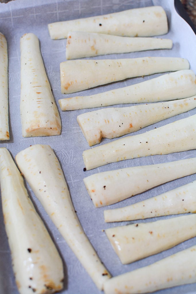 A image of 14 pieces of fresh and peeled parsnips that are cut in half (lengthwise) laid out on a baking sheet on a baking pan
