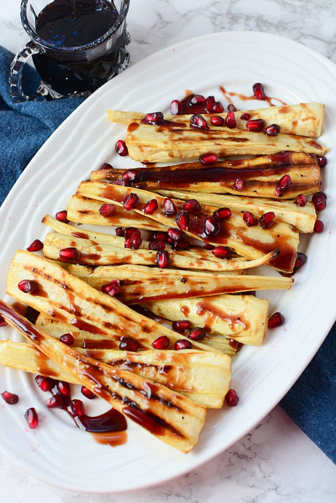 An image of long sliced of oven roasted parsnips drizzled with some honey pomegranate topped with pieces of pomegranate seeds, served on a white platter sitting on a piece of blue cloth. Beside it is a glass of the pomegranate sauce.