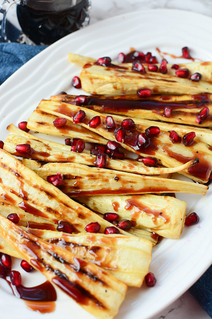 An image of long sliced of oven roasted parsnips drizzled with some honey pomegranate topped with pieces of pomegranate seeds, served on a white platter sitting on a piece of blue cloth.