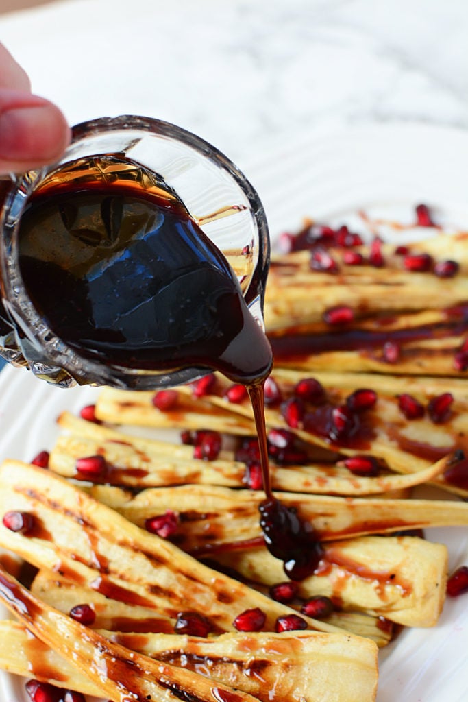 Honey pomegranate juice being poured from a small glass onto pieces of cooked parsnips with pomegranate seeds