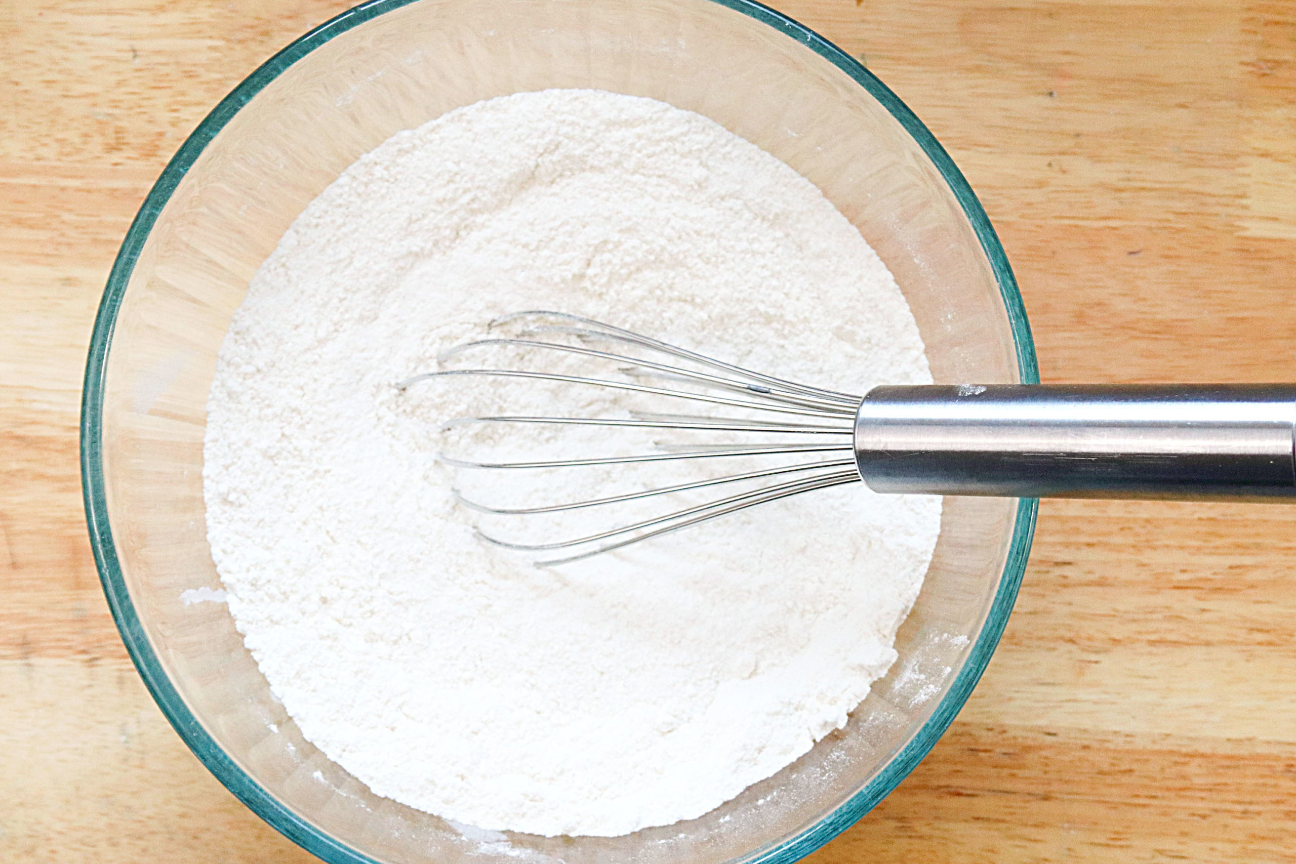 A glass bowl holds the dry ingredients with a whisk resting it in.