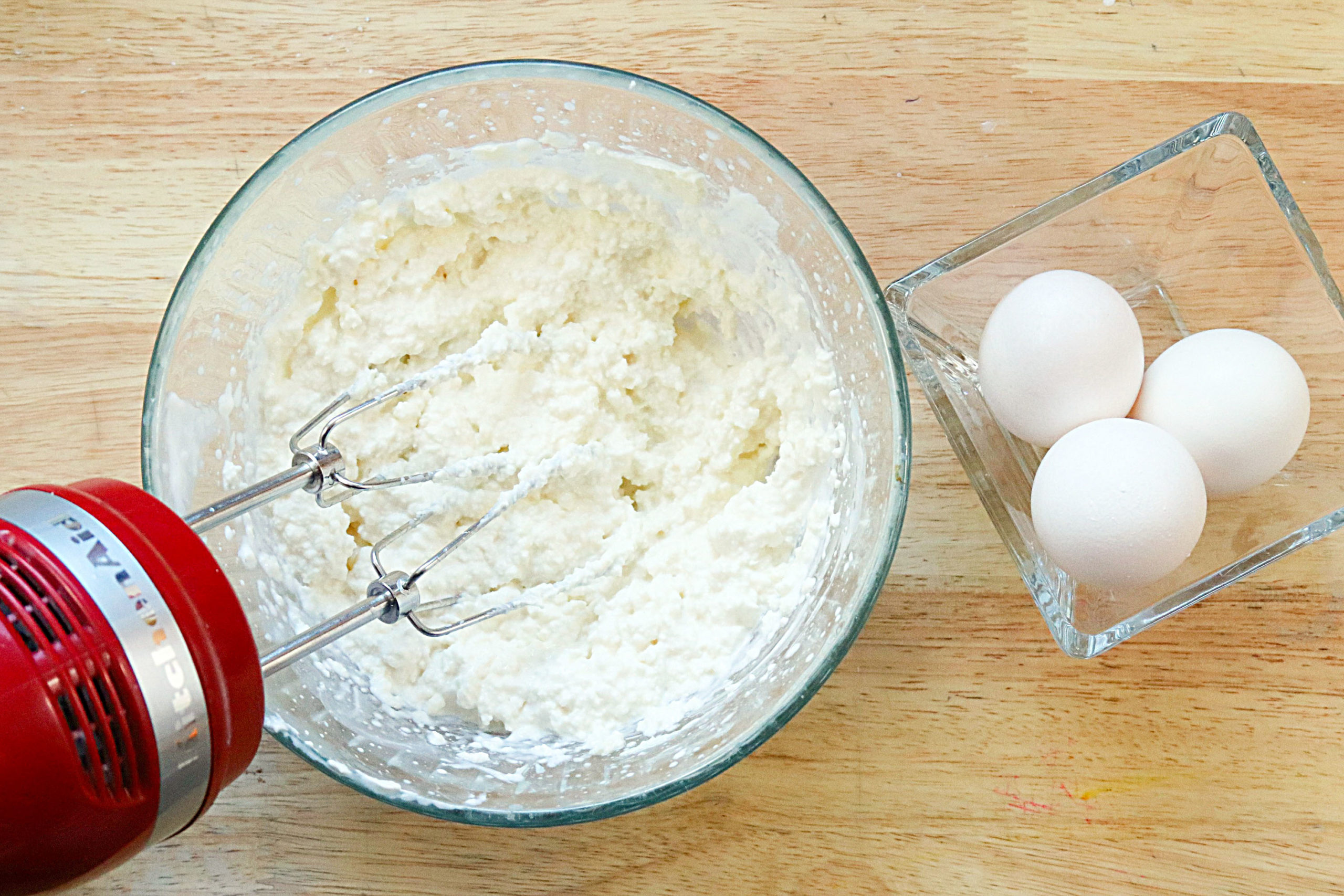 A red handmixer mixes the cake batter. There are three eggs in a glass bowl next to the batter on the right.