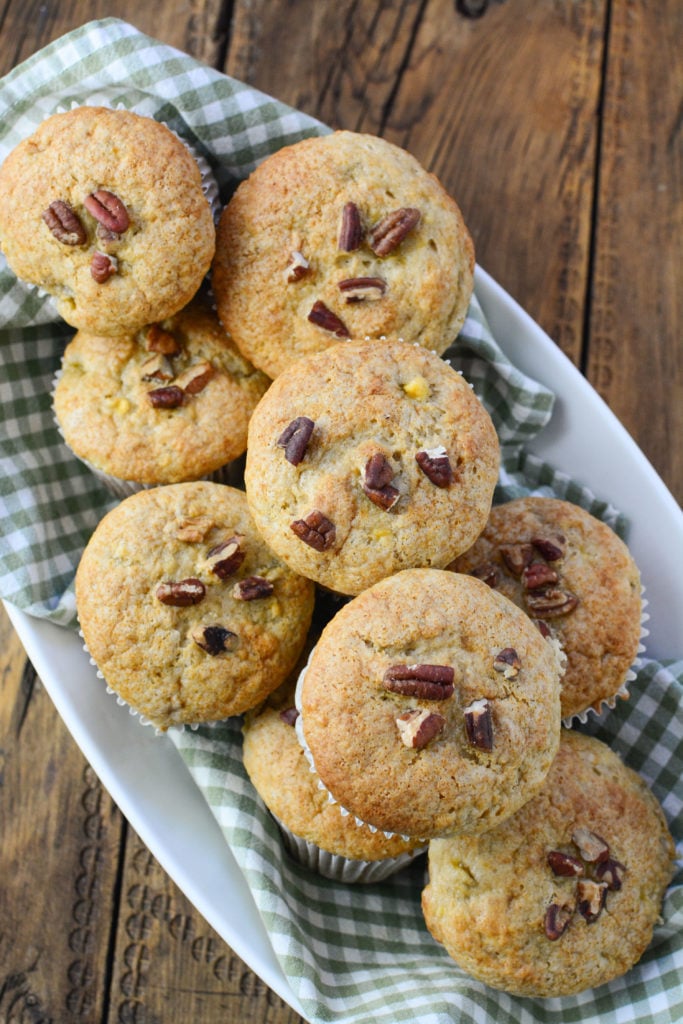 12 banana pecan muffins, with pecans on top, on a white serving plate waiting to pair with a cup of coffee!