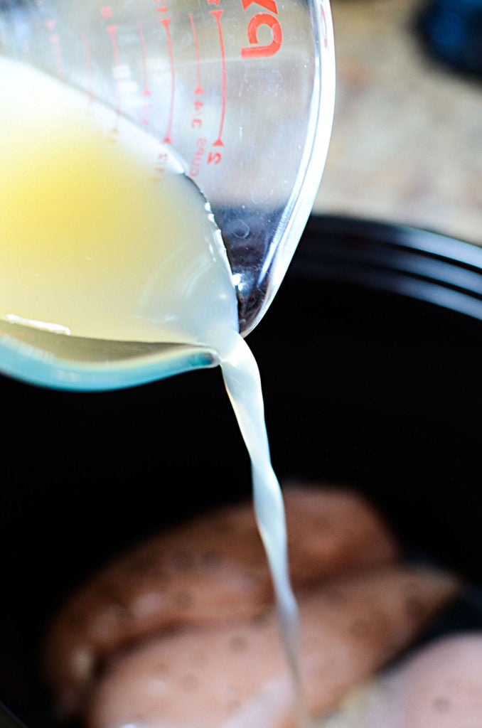 Chicken broth being poured from a measuring cup to a slow cooker with three pieces of boneless and skinless chicken breasts