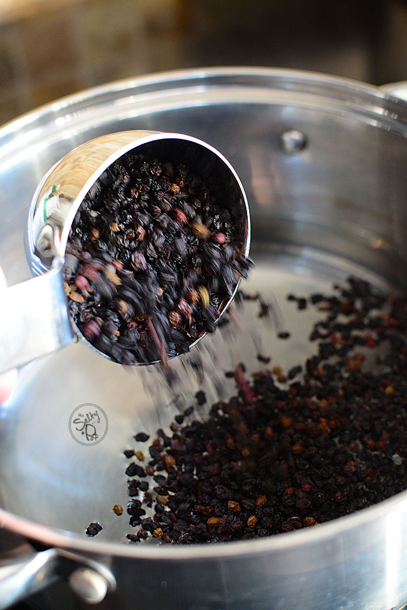 An image of a stainless measuring cup where dried elderberries are being poured into a deep cooking pot