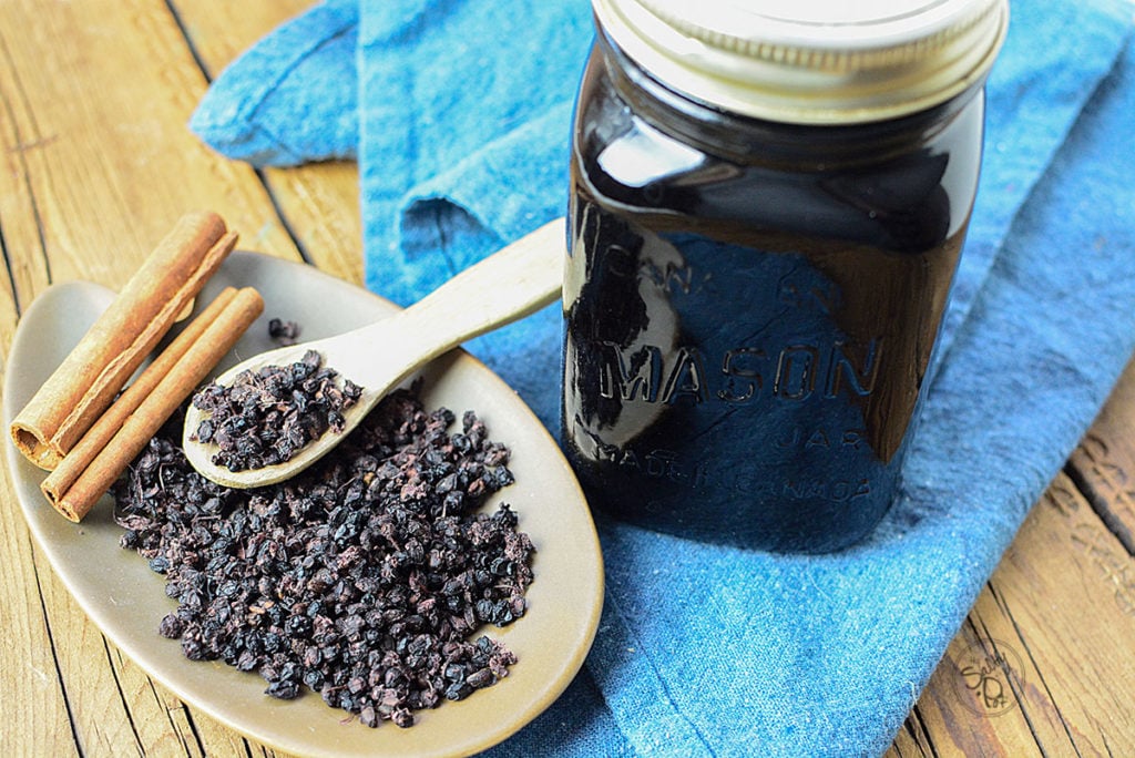 An image of dried elderberry on a brown plate with a wooden spoon with cinnamon sticks on the side. There's also a mason jar full of elderberry syrup with lid, sitting on a piece of blue cloth.