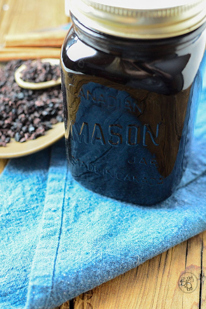 A jar of elderberry honey syrup with lid sitting on a piece of navy blue cloth. Behind it is a plate full of dried elderberries with a wooden spoon on top full of some dried elderberries