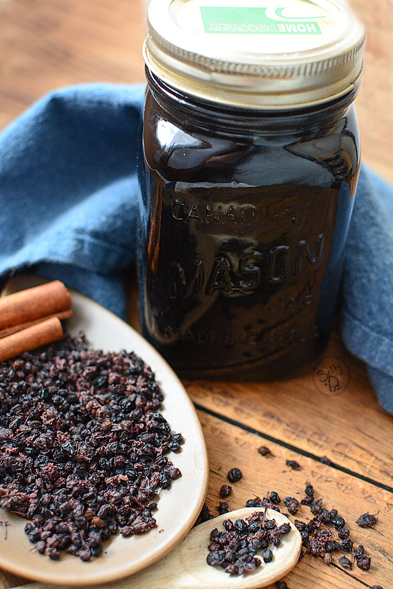A jar of elderberry syrup with lid. Behind the jar is a piece of navy blue cloth. Beside it is plate of dried elderberries with a few cinnamon sticks on the side. Next to it is a wooden spoon with some dried elderberries
