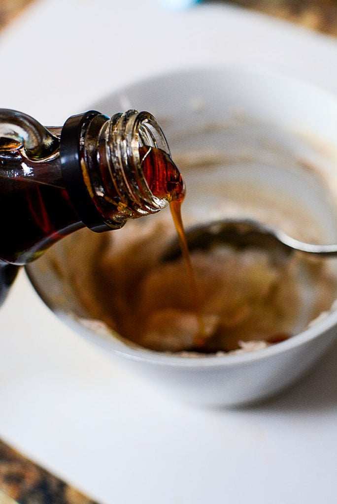 Maple syrup from a bottle is being poured onto the cinnamon and butter mixture in a small bowl with spoon