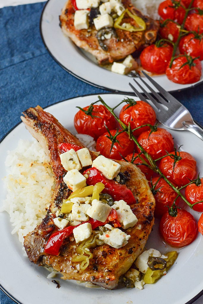 A huge piece of GRILLED MEDITERRANEAN PORK CHOPS topped with pieces of cheese and peppers sitting on steamed rice with ten pieces of roasted vine tomatoes on the side. The dish is served in a white circle plate with a fork on the side of the plate. At the background is another serving of the same dish. The two plates are laid out on a navy blue cloth.