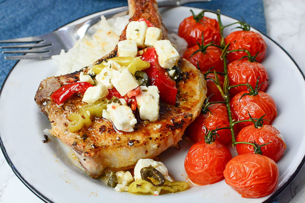 A huge piece of GRILLED MEDITERRANEAN PORK CHOPS topped with pieces of cheese and peppers sitting on steamed rice with ten pieces of roasted vine tomatoes on the side. The dish is served in a white circle plate with a fork on the side of the plate. The plate is sitting on a navy blue cloth.