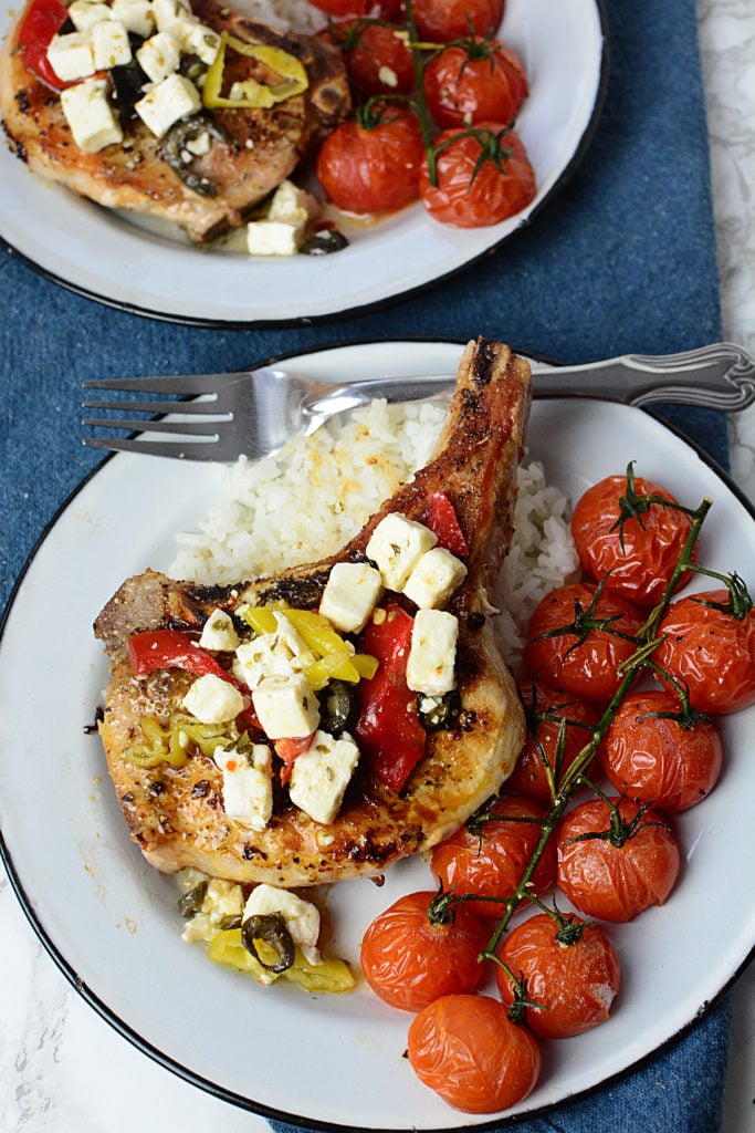 A huge piece of GRILLED MEDITERRANEAN PORK CHOPS topped with pieces of cheese and peppers sitting on steamed rice with ten pieces of roasted vine tomatoes on the side. The dish is served in a white circle plate with a fork on the side of the plate. At the background is another serving of the same dish. The two plates are laid out on a navy blue cloth.