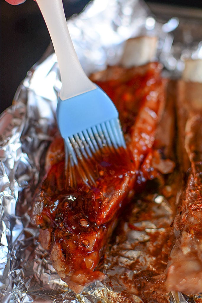 Two pieces of ribs on a foil being brushed with BBQ sauce using a blue brush