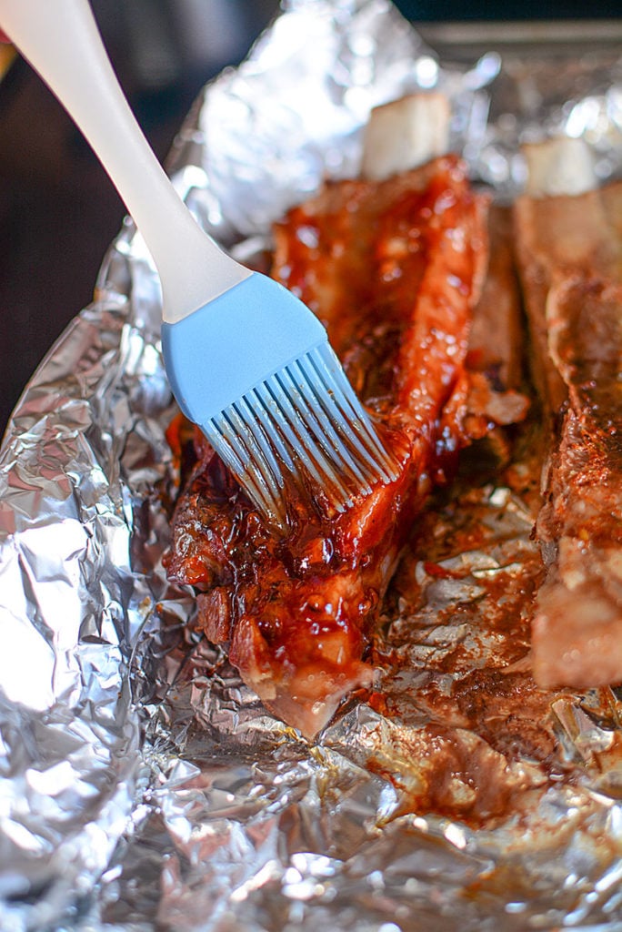 Two pieces of ribs on a foil being brushed with BBQ sauce using a blue brush