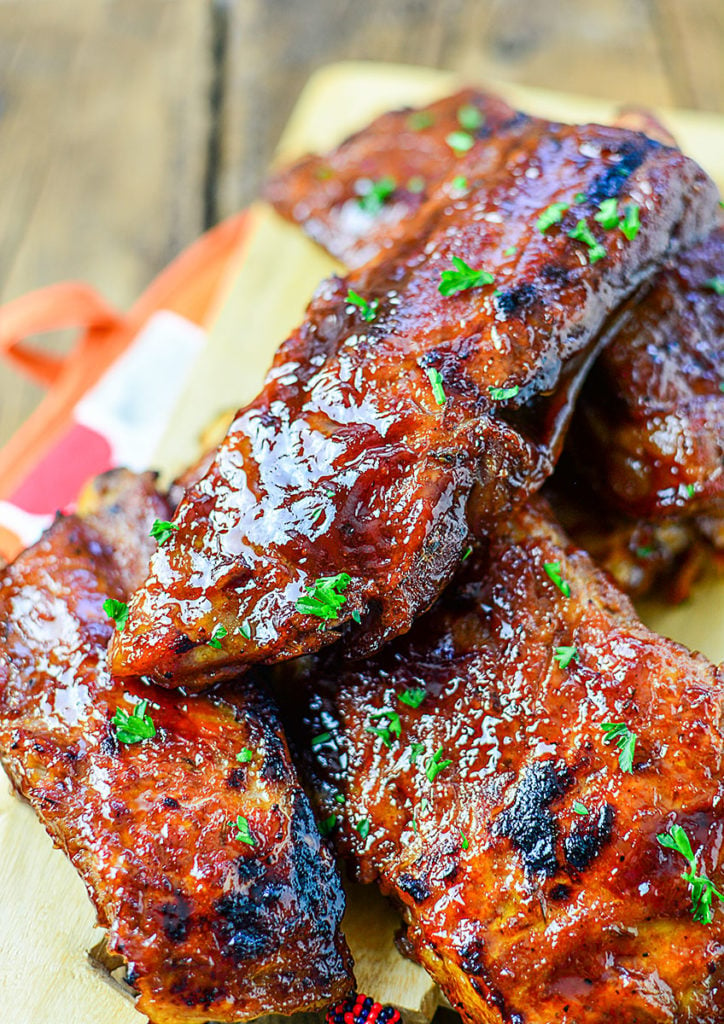 Close up image of 5 pieces of OVEN ROASTED LOW AND SLOW BBQ RIBS sitting on a wooden chopping board, garnished with some chopped parsley.